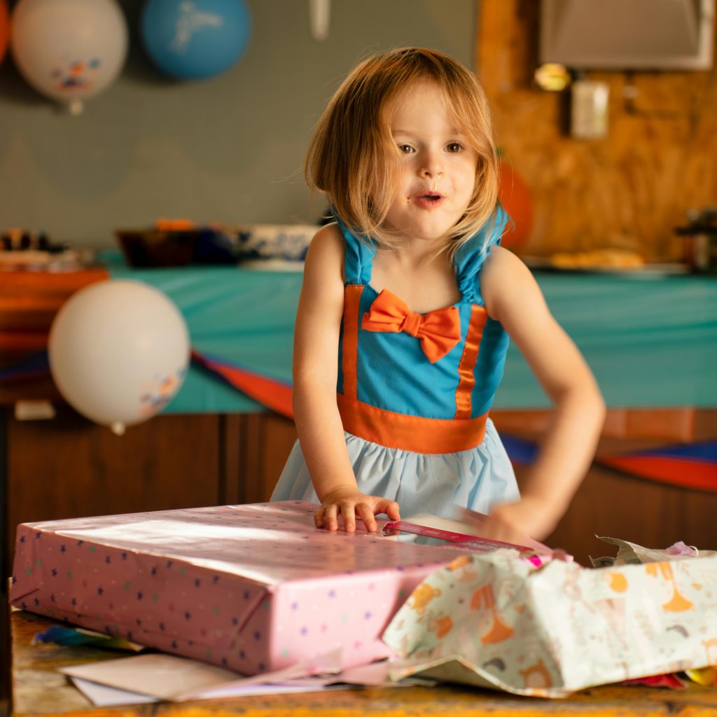 Young girl excitedly opening a gift box during her birthday celebration indoors.